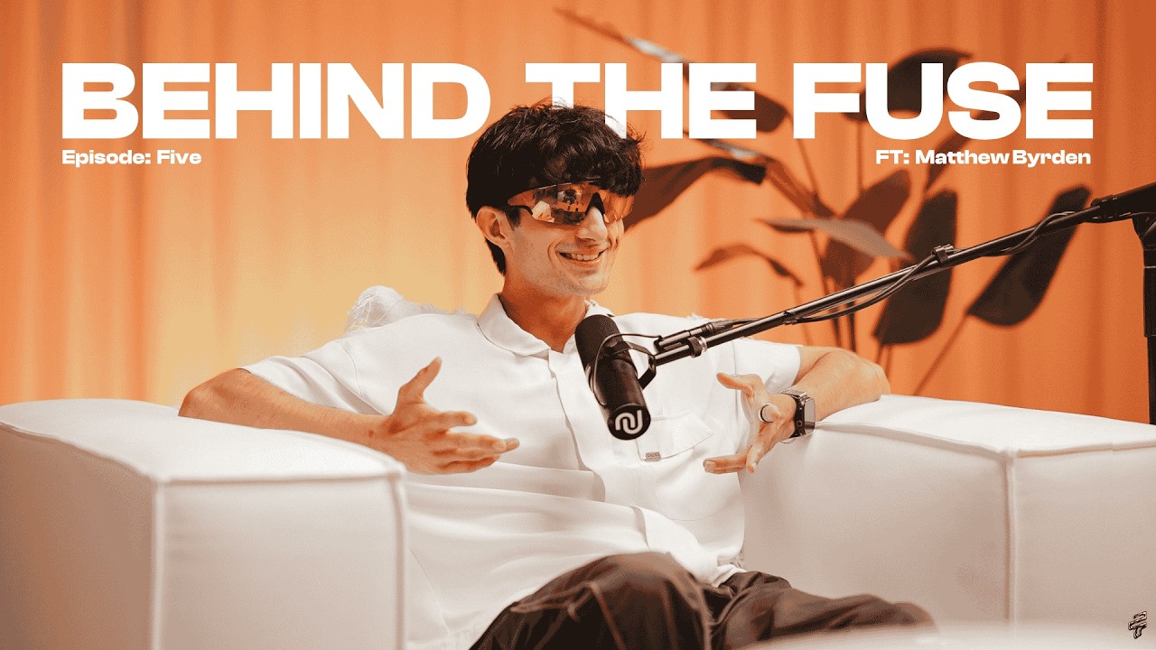 Matthew Byrden on the Behind The Fuse podcast set, laughing mid-gesture on a white couch in front of an orange curtain